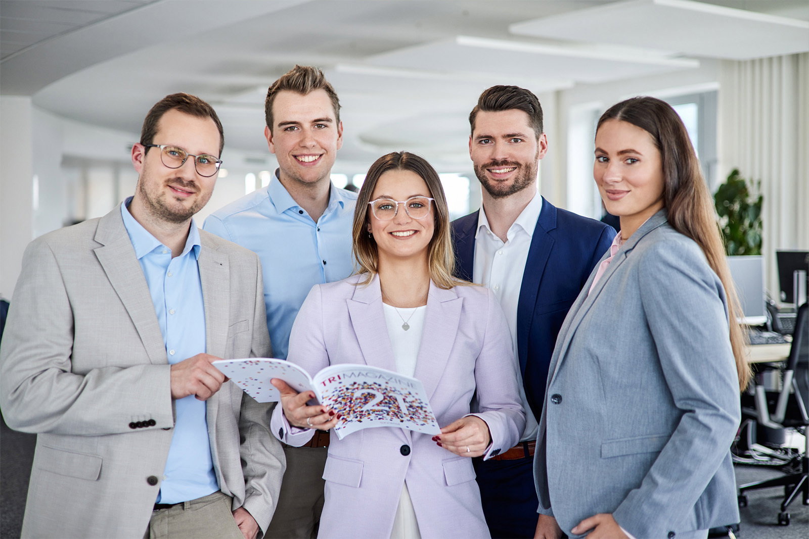 Fünf Consultants von TriFinance blicken im Düsseldorfer Büro lächelnd in die Kamera und vertreten unsere Finance-Leistungen.