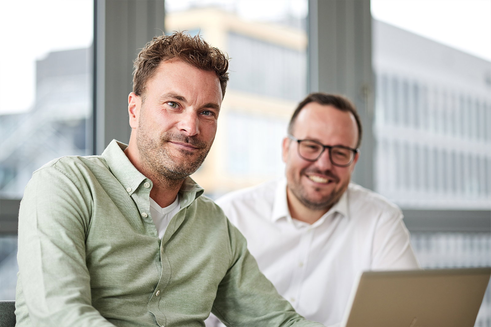 TriFinance Senior Berater-Team in Düsseldorf blickt lächelnd in die Kamera, ein Laptop vor ihnen für strategisches Controlling & Management Reporting.