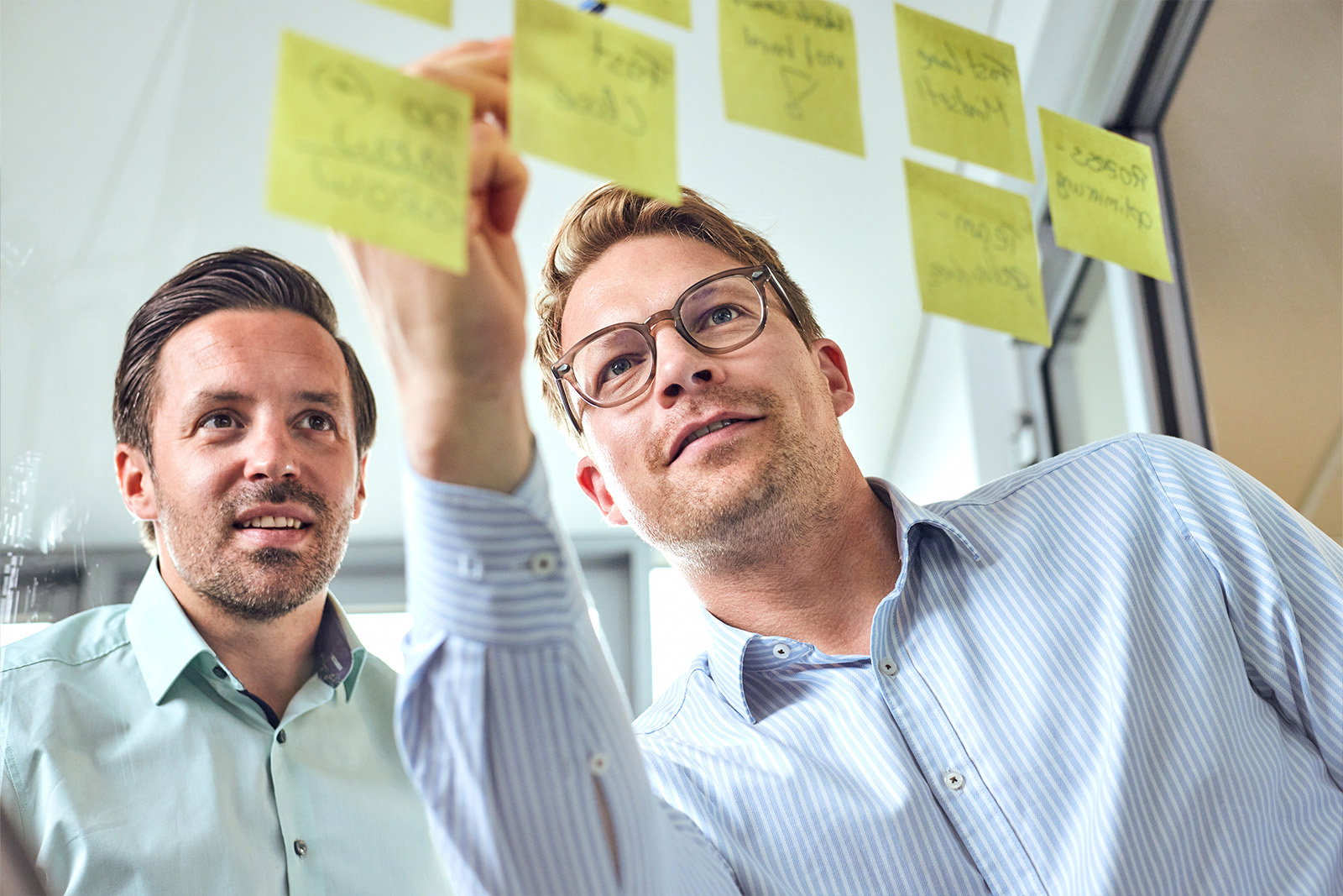 Zwei TriFinance Berater in Frankfurt planen an einer Glaswand mit gelben Haftnotizen eine ERP-Implementierung und Transformation.