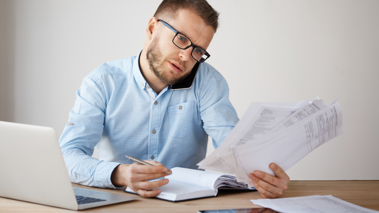Busy concentrated unshaved businessman in glasses and shirt sitting in a comfortable light office, looking through papers, planning meeting with a team of workers by phone.