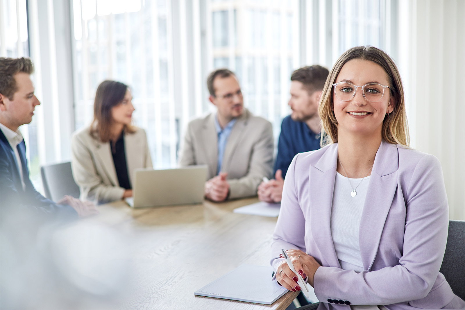 TriFinance Senior Project Consultant mit Brille im fliederfarbenen Blazer lächelt in Düsseldorf in die Kamera, ihr Berater-Team bespricht Leistungen dahinter.