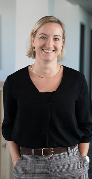 Die Recruiterin Marie von TriFinance Deutschland steht im Office in schwarzem Top und lächelt in die Kamera, ihre Hände lässig in den Hosentaschen.